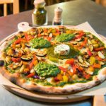Une pizza garnie de légumes rôtis colorés et de pesto, servie sur une planche en bois.