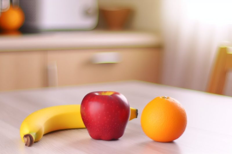 Trois fruits différents (par exemple une pomme, une banane et une orange) disposés sur une table en bois claire.