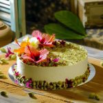 Un gâteau délicat décoré de fleurs d'hibiscus et de graines de cardamome posé sur une table rustique.